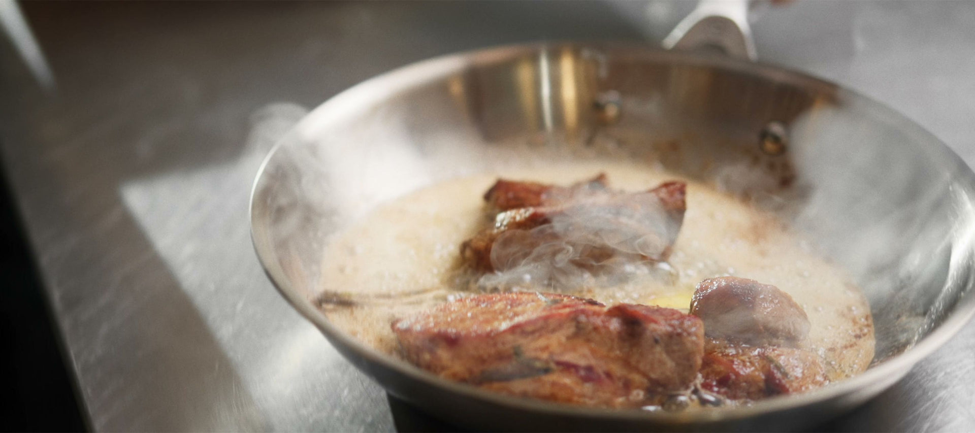 En stekpanna i rostfritt stål där kött steks i skummande smör medan ångan stiger