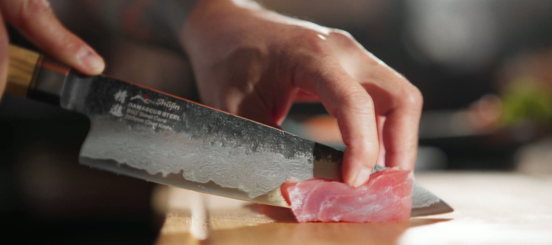 En hand skär en bit tonfisk med en Shōjin-kniv med damaskusmönster på en skärbräda i trä