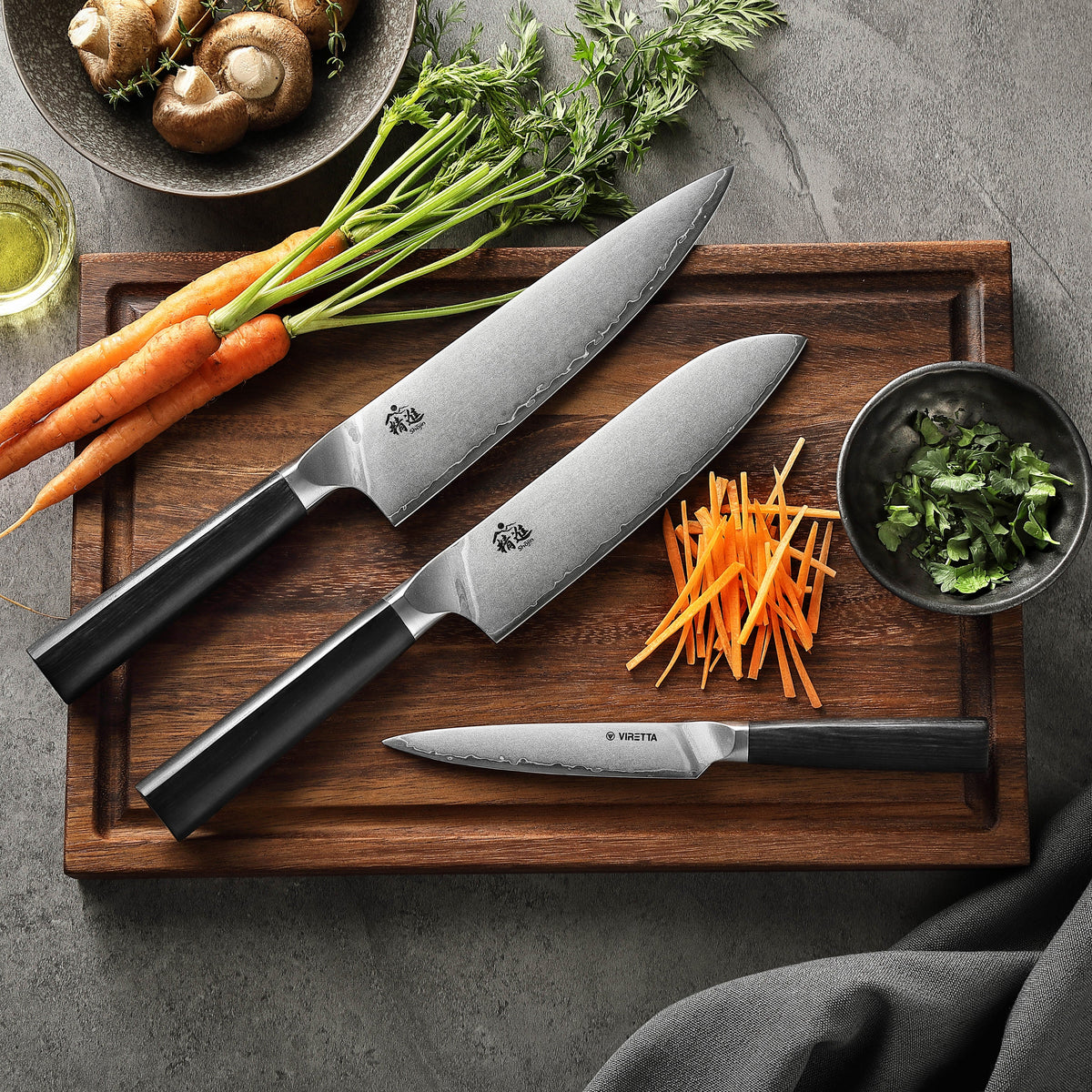 Shōjin Aira santokukniv, kockkniv och universalkniv på en skärbräda i valnöt med morötter