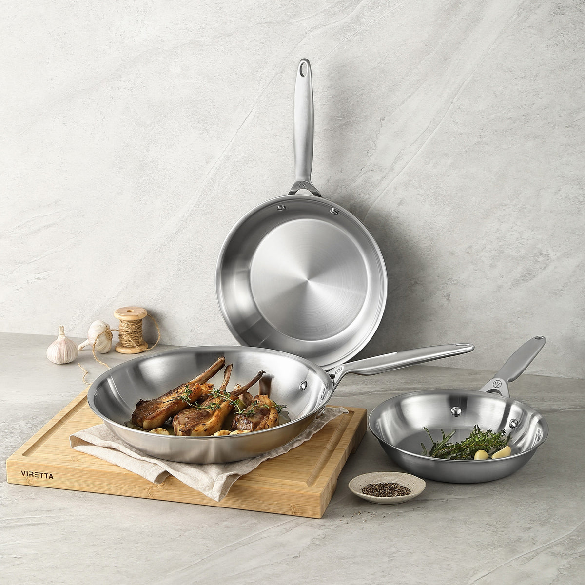 Tre stekpannor i 3-Ply rostfritt stål, den främsta med lammkotletter på en skärbräda