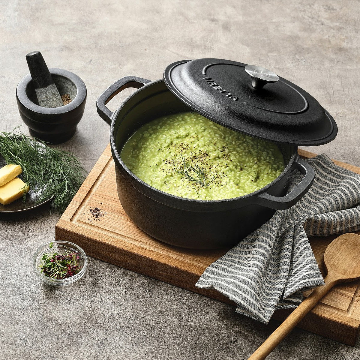Skiffersvart gjutjärnsgryta med grön risotto och locket på glänt, på en skärbräda