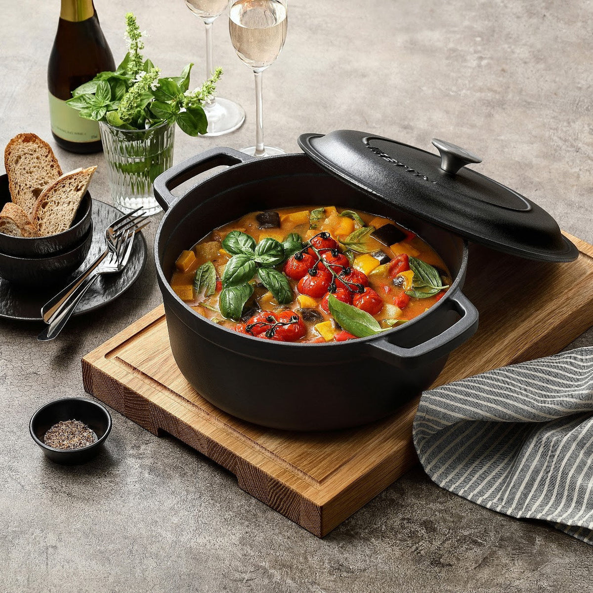 Skiffersvart gjutjärnsgryta med ratatouille och locket på glänt, på en skärbräda