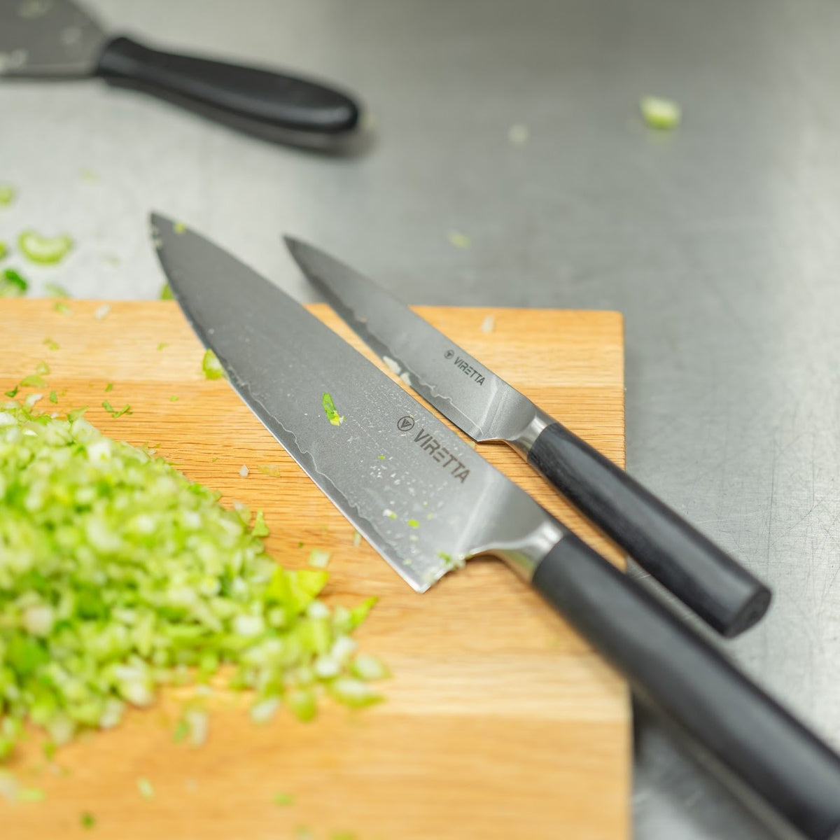 Shōjin Aira kockkniv och en mindre Aira-kniv på en skärbräda i ek med hackad blekselleri