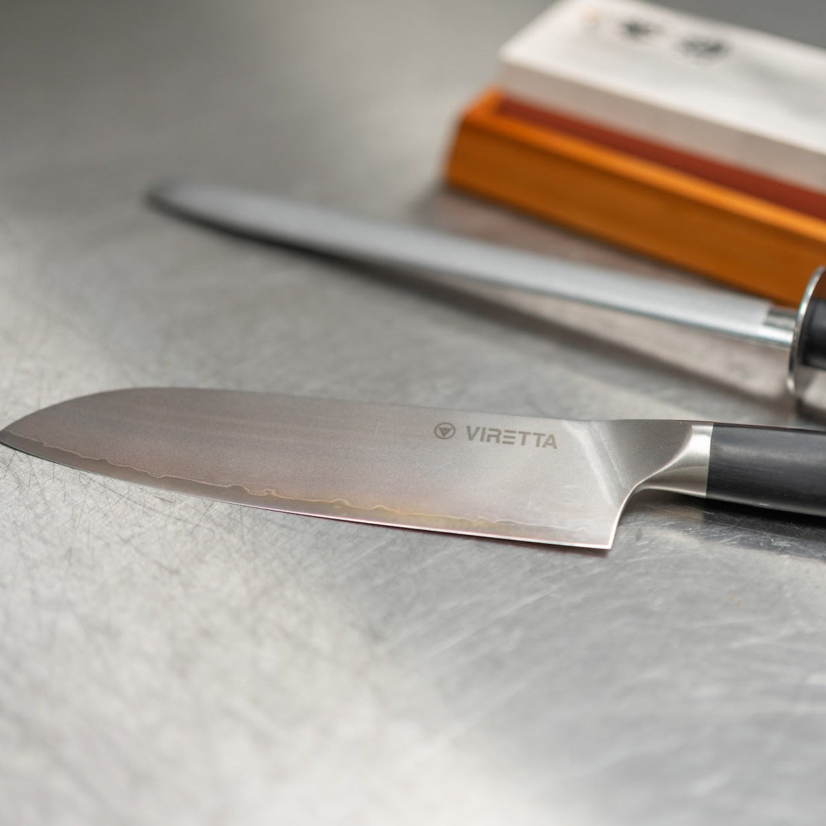 Shōjin Aira santokukniv framför ett skärpstål och en slipsten i bambuhållare på en rostfri bänk
