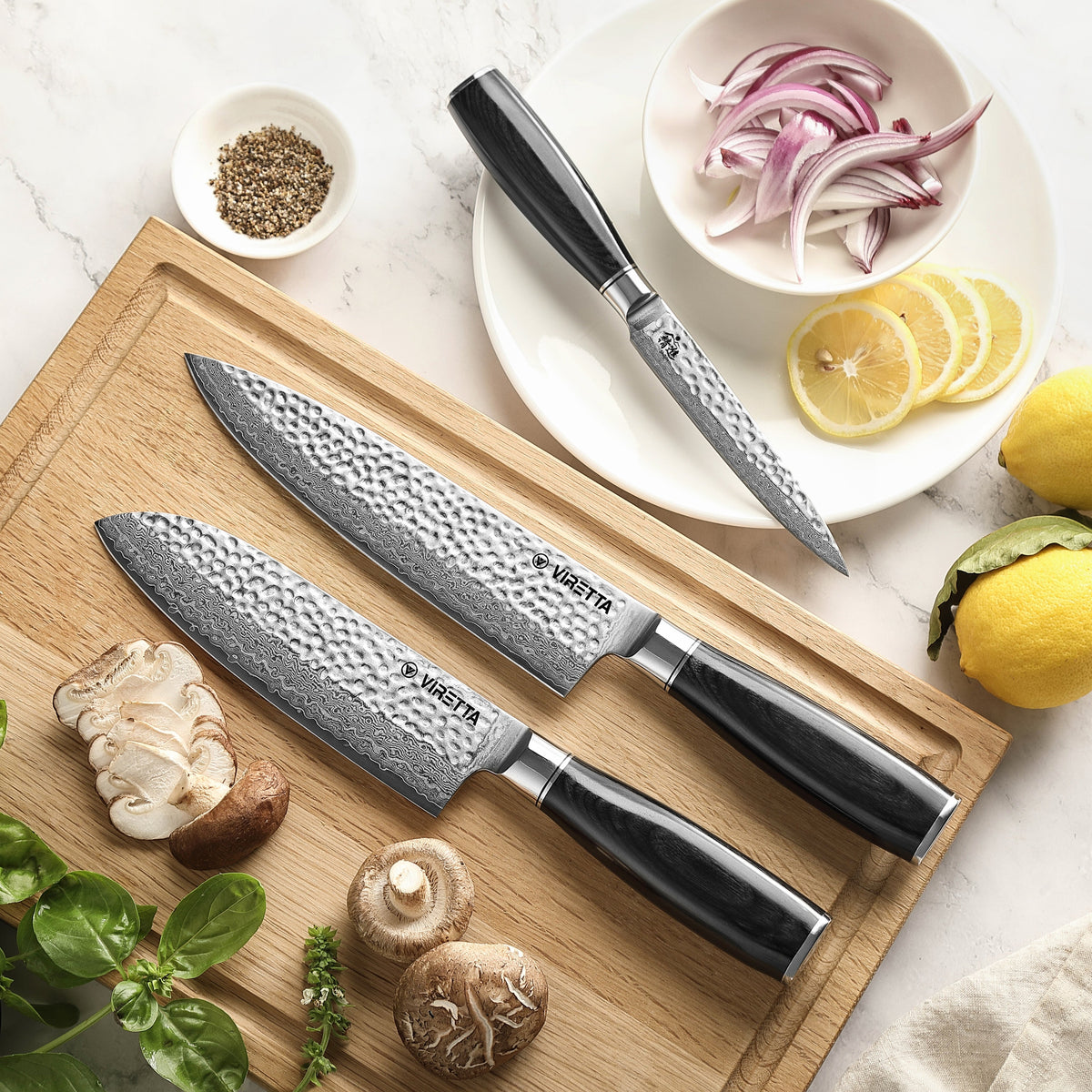 Shōjin Taru santokukniv och kockkniv på en skärbräda i ek, universalkniv bredvid rödlök och citron