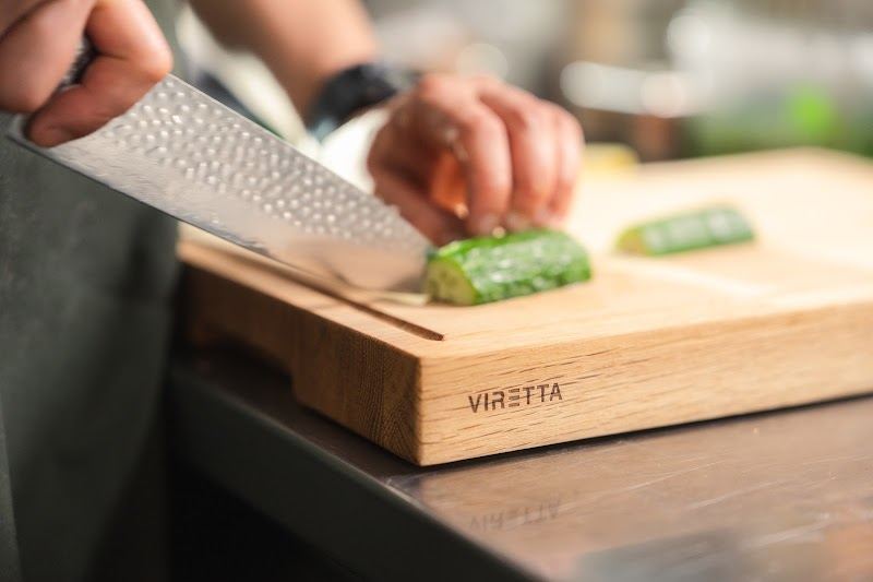 En kniv med hamrat blad skär gurka på en skärbräda i ek. Brädan har Viretta-logotypen