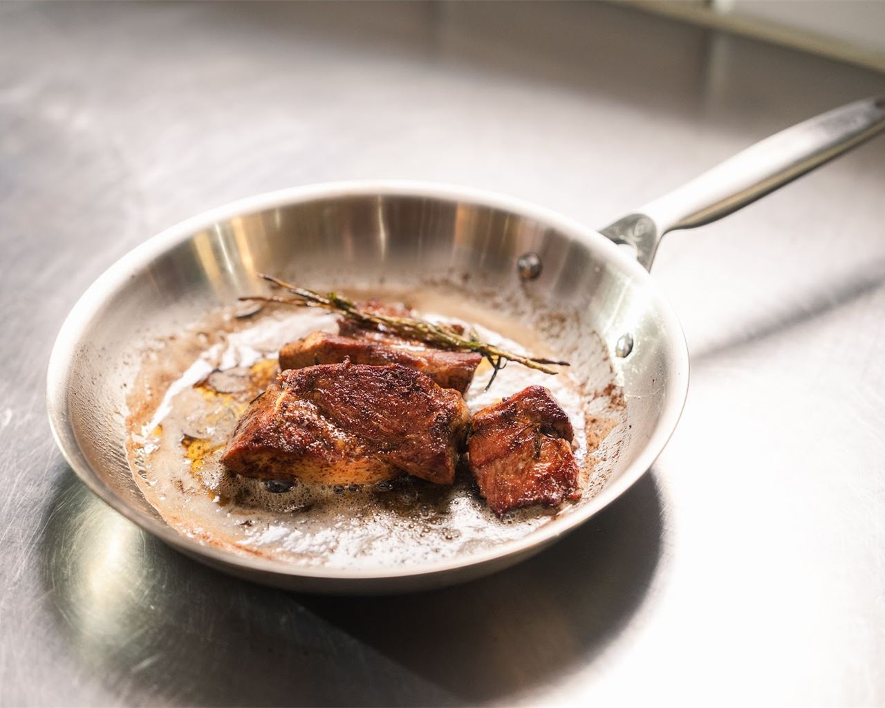 En stekpanna i rostfritt stål med stekt kött och en örtkvist på ett bord av stål
