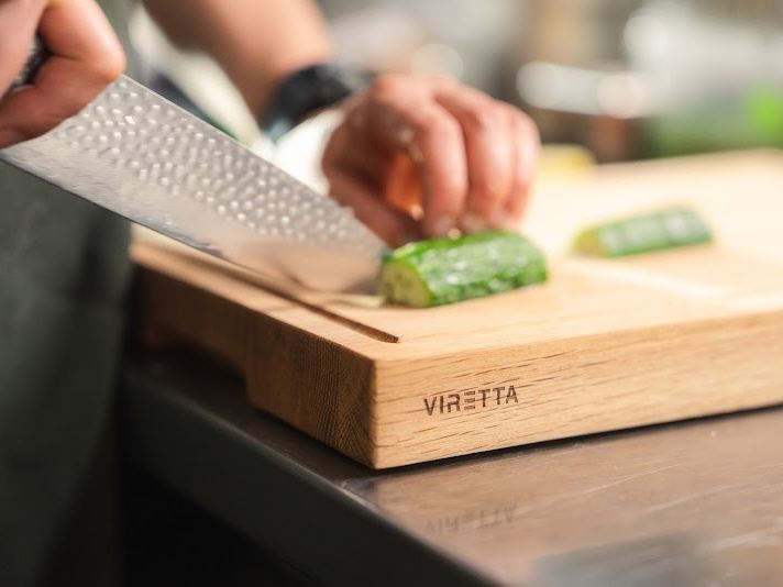 En kniv med hamrat blad skär gurka på en skärbräda i ek. Brädan har Viretta-logotypen