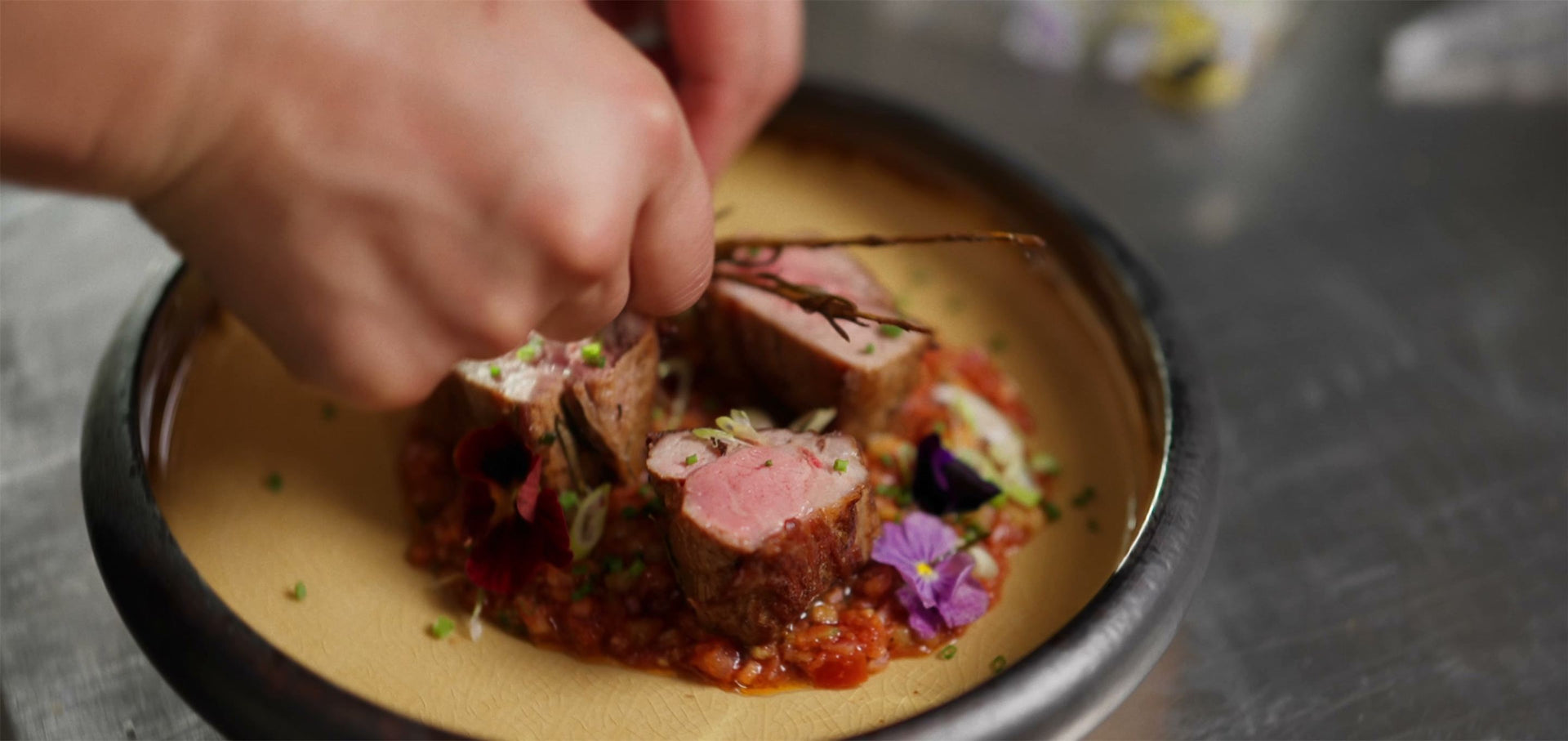 En hand lägger sista handen vid en uppläggning med rosa kött, tomat och ätliga blommor på en tallrik med mörk kant