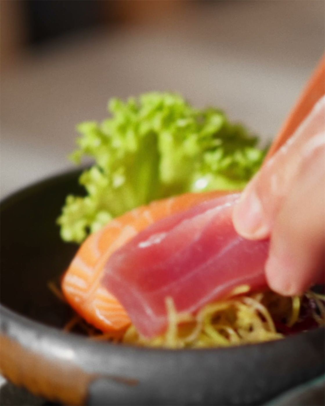 En hand lägger en bit tonfisk på en mörk tallrik med lax och sallad