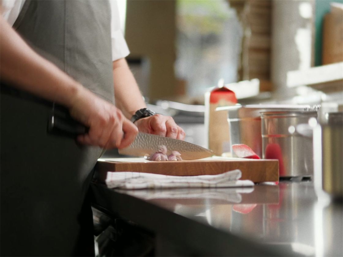 En kock i grått förkläde hackar schalottenlök med en kniv med hamrat blad på en skärbräda i ett restaurangkök
