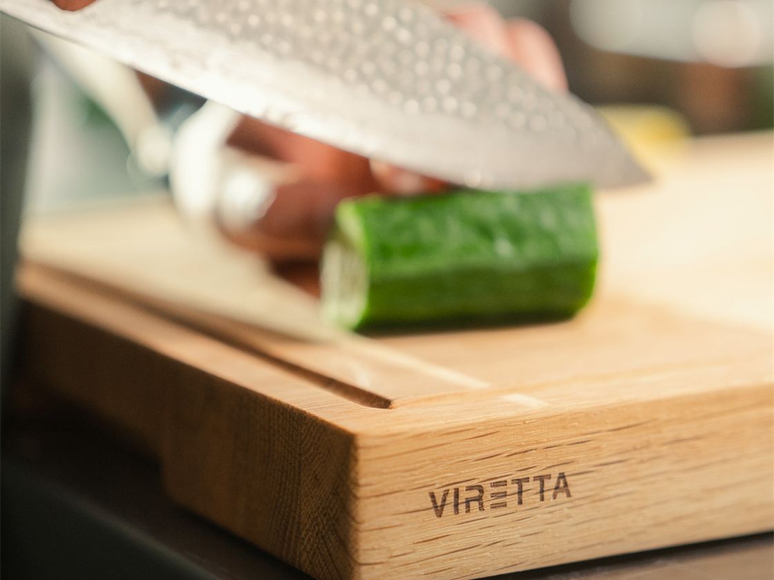 En kniv skär gurka på en skärbräda i trä. Skärbrädan har Viretta-logotypen